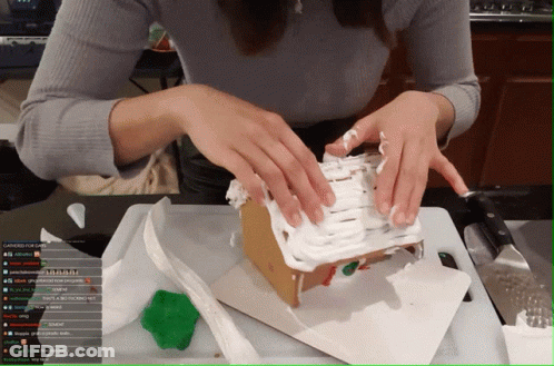 A Woman Is Decorating A Gingerbread House On A Cutting Board While Watching A Stream On Youtube GIF