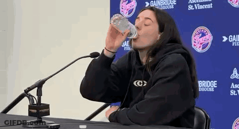 A Woman Is Drinking Water From A Bottle While Sitting At A Table With A Microphone . GIF