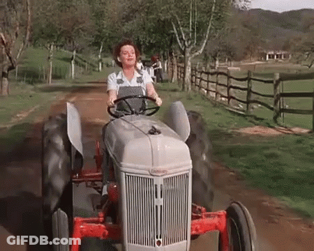A Woman Is Driving A Tractor Down A Dirt Road In A Field . GIF