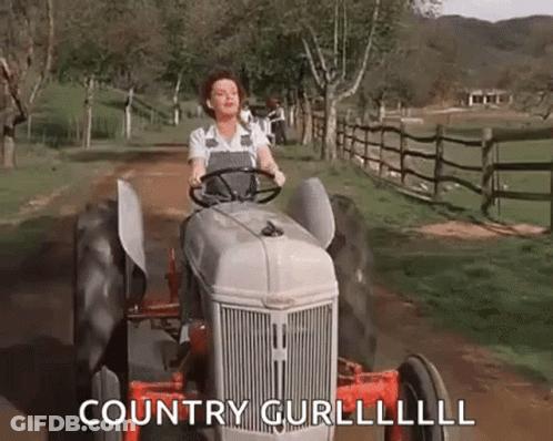 A Woman Is Driving A Tractor Down A Dirt Road In A Field . GIF