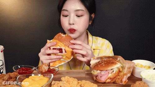 A Woman Is Eating A Hamburger And Fried Chicken On A Table . GIF