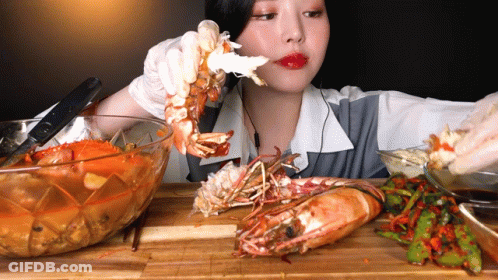 A Woman Is Eating A Large Shrimp From A Bowl On A Cutting Board GIF
