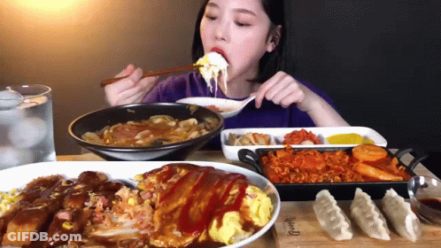 A Woman Is Eating A Plate Of Food With A Spoon And Chopsticks While Sitting At A Table GIF