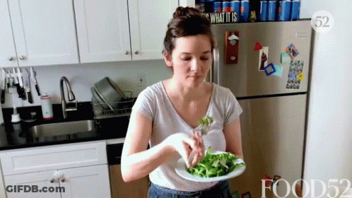 A Woman Is Eating A Salad In A Kitchen With A Sign That Says What It Is On The Refrigerator GIF
