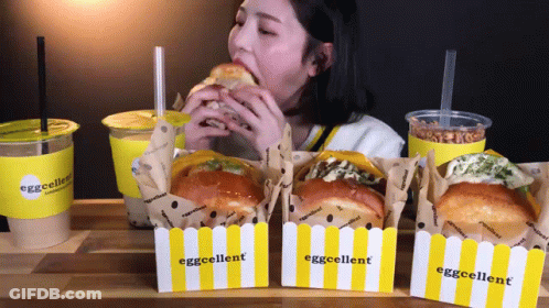 A Woman Is Eating A Sandwich In Front Of Boxes That Say Eggcellent On Them GIF