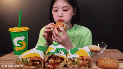 A Woman Is Eating A Subway Sandwich Next To A Cup That Says ' S ' On It GIF
