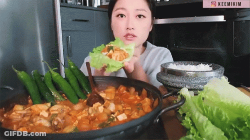 A Woman Is Eating A Wrap Of Lettuce In Front Of A Pot Of Food And A Sign That Says Keemikim GIF