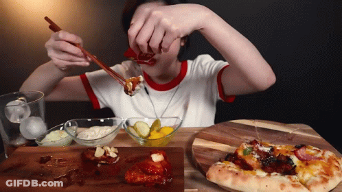 A Woman Is Eating Food With Chopsticks And A Pizza On A Wooden Cutting Board GIF