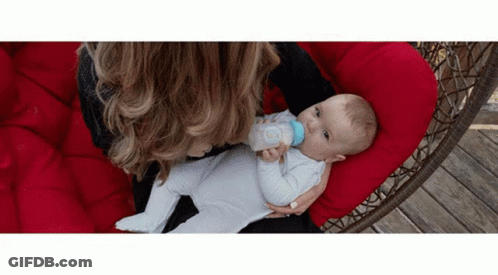 A Woman Is Feeding A Baby From A Bottle While Sitting On A Red Chair . GIF