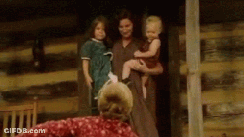A Woman Is Holding A Baby And A Dog In Her Arms While Standing Next To A Log Cabin . GIF