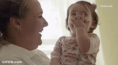 A Woman Is Holding A Baby Girl In Her Arms And The Baby Is Covering Her Face With Her Hands . GIF