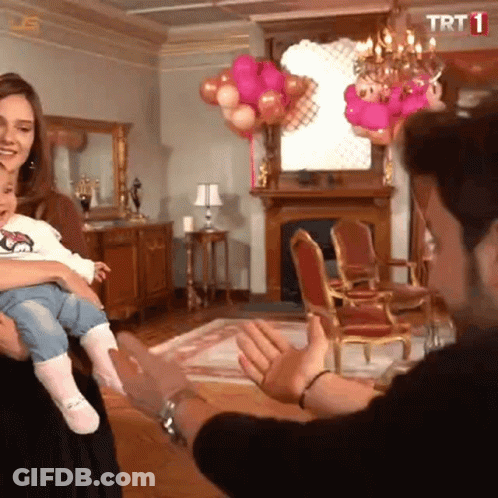 A Woman Is Holding A Baby In A Living Room With Balloons And A Trt Logo GIF