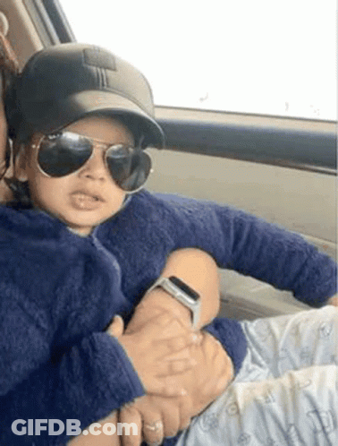 A Woman Is Holding A Baby In Her Arms In A Car . The Baby Is Wearing Sunglasses And A Baseball Cap . GIF