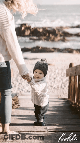 A Woman Is Holding A Baby 'S Hand While They Walk On A Boardwalk GIF
