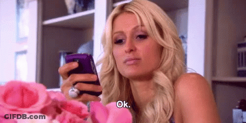 A Woman Is Holding A Cell Phone In Her Hand While Sitting In Front Of A Bouquet Of Pink Roses . GIF