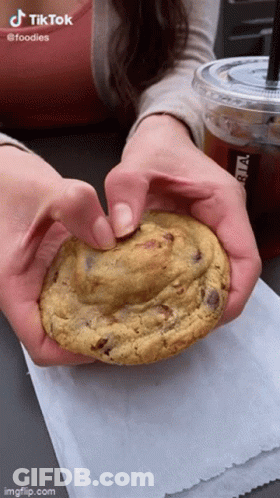A Woman Is Holding A Cookie In Her Hands With A Cup Of Coffee In The Background . GIF