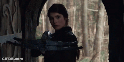 A Woman Is Holding A Crossbow In Front Of A Door In A Forest . GIF