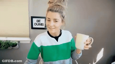 A Woman Is Holding A Cup In Front Of A Sign That Says Be Dumb . GIF