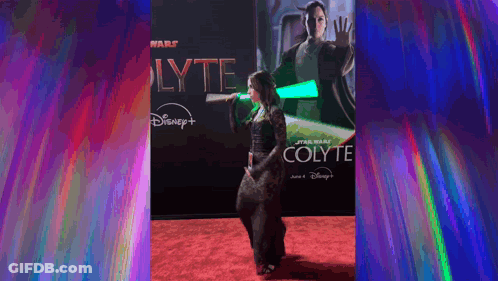 A Woman Is Holding A Green Lightsaber On A Red Carpet In Front Of A Poster For Star Wars Colyte . GIF