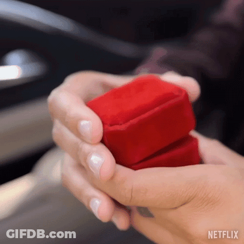 A Woman Is Holding A Red Box With A Ring Inside Of It . GIF