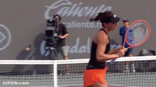 A Woman Is Holding A Tennis Racquet On A Court In Front Of A Wall That Says Caliente GIF