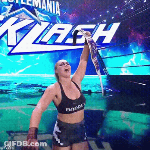 A Woman Is Holding A Wrestling Championship Belt In The Air While Standing On A Stage . GIF