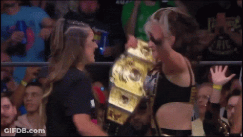 A Woman Is Holding A Wrestling Championship While Another Woman Watches GIF