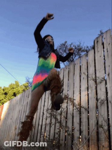 A Woman Is Jumping Over A Wooden Fence Wearing A Colorful Dress GIF