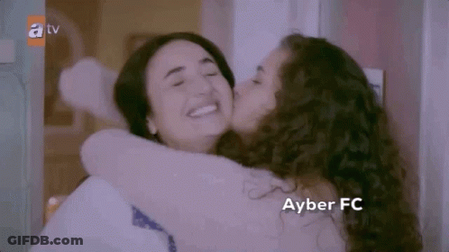 A Woman Is Kissing Another Woman On The Cheek While Hugging Each Other . GIF