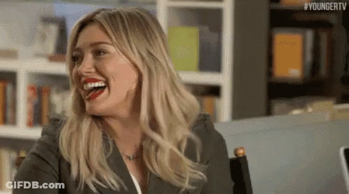 A Woman Is Laughing While Sitting At A Table In Front Of A Bookshelf . GIF