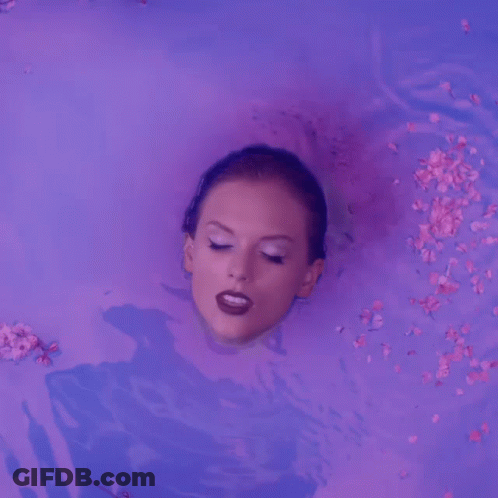 A Woman Is Laying In A Bathtub With Flowers Falling Around Her GIF