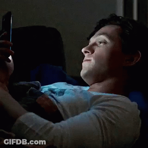 A Woman Is Laying In Bed Looking At Her Phone And Smiling . GIF