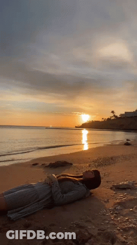 A Woman Is Laying On A Beach With The Sun Setting In The Background GIF