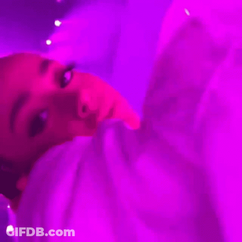 A Woman Is Laying On A Bed Under A Purple Light And Looking At The Camera . GIF