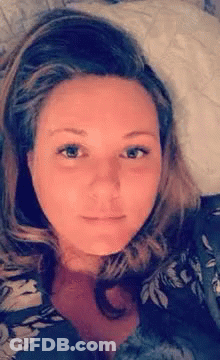 A Woman Is Laying On A Bed With A Pillow In Her Lap And Looking At The Camera . GIF