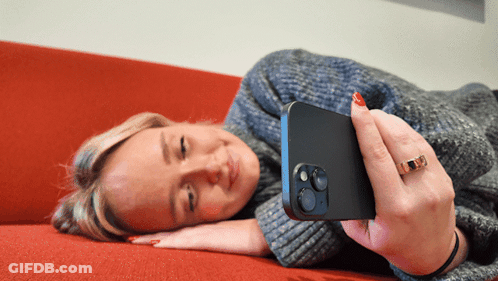 A Woman Is Laying On A Red Couch Looking At Her Cell Phone GIF