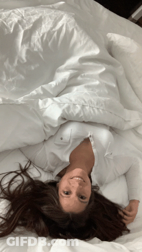 A Woman Is Laying On Her Back On A Bed With White Sheets GIF