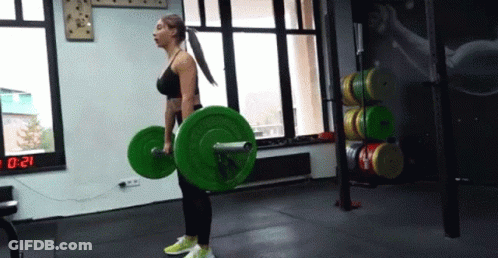 A Woman Is Lifting A Barbell In A Gym With A Digital Clock In The Background . GIF