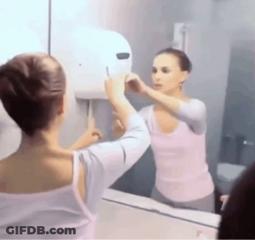 A Woman Is Looking At Her Reflection In A Bathroom Mirror . GIF