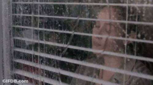 A Woman Is Looking Out A Window With Blinds On It . GIF