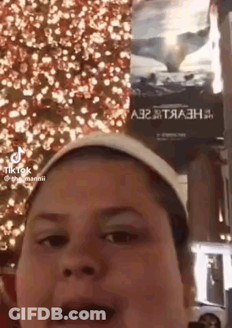 A Woman Is Making A Funny Face In Front Of A Christmas Tree And A Sign That Says A32 GIF