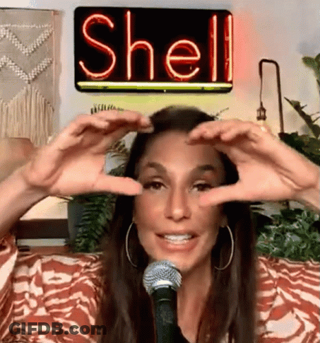 A Woman Is Making A Heart Shape With Her Hands In Front Of A Neon Sign That Says Shell On It . GIF