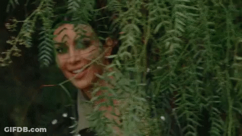 A Woman Is Peeking Out From Behind A Tree And Smiling At The Camera . GIF