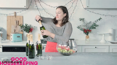 A Woman Is Pouring A Bottle Of Champagne In A Kitchen With The Words Good Housekeeping On The Bottom GIF