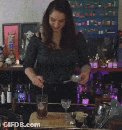 A Woman Is Pouring A Drink Into A Glass In Front Of A Shelf Of Bottles GIF