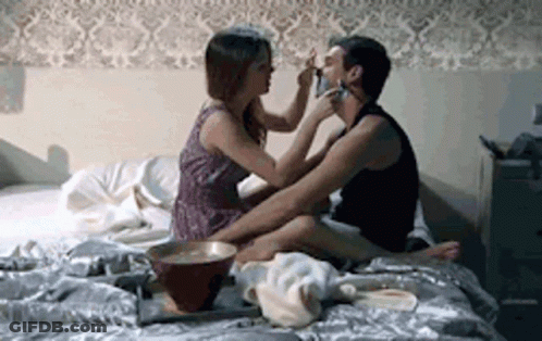 A Woman Is Putting Makeup On A Man 'S Face While They Are Sitting On A Bed . GIF