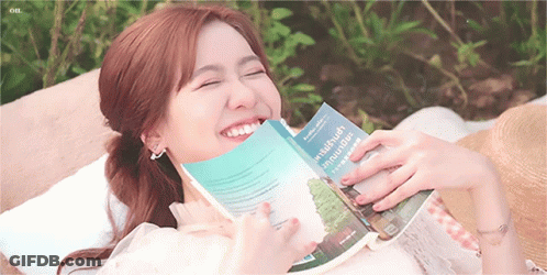 A Woman Is Reading A Book And Smiling While Laying On A Blanket GIF