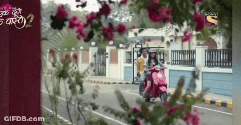 A Woman Is Riding A Scooter Down A Street With Flowers In The Foreground And A Sony Channel Logo In The Background GIF