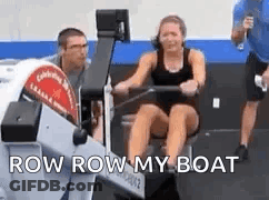 A Woman Is Rowing A Boat In A Gym With A Man Standing Behind Her . GIF