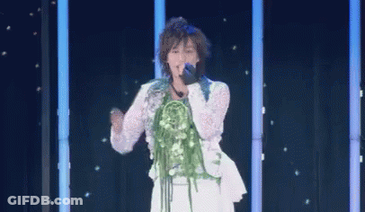 A Woman Is Singing Into A Microphone On A Stage With A Bouquet Of Flowers On Her Neck . GIF
